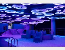 Ein Innenraum mit futuristischem Design, beleuchtet durch zahlreiche kreisförmige LED-Lichter an der Decke in Violett- und Blautönen. Der Raum enthält Rutschen und einen Pool mit weißen Kunststoffbällen, die eine spielerische Atmosphäre erzeugen.