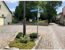 Kreuzung einer gepflasterten Straße mit Straßenschildern „Dr.-Wagner-Weg“ und „Martinistraße“, umgeben von niedrigen Wohnhäusern mit roten Dächern, einem kleinen bepflanzten Bereich und Bäumen im Hintergrund.