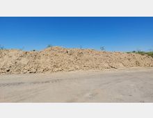 Ein großer Erd- oder Sandhügel auf einer offenen, unbefestigten Fläche, umgeben von klarem, blauem Himmel und vereinzelter Vegetation im Hintergrund.
