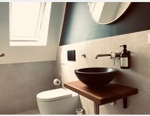 Modernes Badezimmer mit einem schrägen Dachfenster, einer wandmontierten Toilette und einem minimalistischen Waschbereich. Das Waschbecken besteht aus dunklem Stein und ist auf einer hölzernen Ablage montiert, ergänzt durch einen schlichten Wand-Wasserhahn und einen Seifenspender.