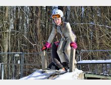 Ein Kind in einem Skianzug mit Helm und Skibrille steht auf einer schneebedeckten Holzrampe vor einem Hintergrund aus kahlen, winterlichen Bäumen.