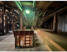 Innenansicht eines historischen Dachbodens mit Holzstruktur. Der Raum zeigt sichtbare Balken, einen Holzfußboden, eine Holztreppe und alte mechanische Geräte, mit einer beleuchteten Notausgangsanzeige an der Decke.