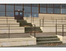 Außenansicht eines Gebäudeeingangs mit breiten Treppenstufen aus Beton, umgeben von beigefarbenen Mauern und Metallgeländern. Im Hintergrund große Fensterfronten mit blickdichtem Glas und einer zentralen Holztür.