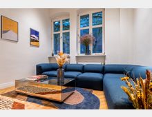 Wohnzimmer mit blauer Eckcouch, Spiegel-Couchtisch und dekorativem Teppich mit geometrischem Muster. Im Hintergrund drei große Fenster mit Blick nach draußen, geschmückt mit Blumen und Dekorationsobjekten auf den Fensterbänken.