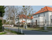 Straßenszene in einem Wohngebiet mit freistehenden weißen Häusern im klassischen Stil und roten Ziegeldächern, umgeben von Zäunen und Hecken. Ein Straßenschild mit der Aufschrift „Wichernstraße“ steht neben einem Gehweg, unter kahlen Bäumen.