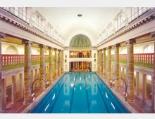 Historisches Hallenbad mit einem langen, rechteckigen Schwimmbecken, umgeben von Säulen und hohen Wänden mit roten Gittern. Der Raum hat eine gewölbte Decke und ist mit Arkaden sowie einer verzierten Kuppel im Hintergrund gestaltet.