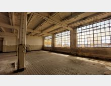 Verlassener Industriebereich mit hohen Decken, großen rechteckigen Fenstern mit vergilbtem Glas und sichtbaren Metallrahmen. Der Raum zeigt abgenutzte Holzdielen, abgeblätterte Farbe an den Wänden und freiliegende Leitungen an der Wand.