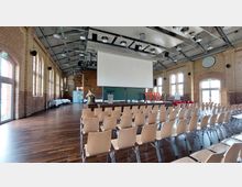 Ein Veranstaltungsraum mit Backsteinwänden, großen Fenstern und einem Holzboden, ausgestattet mit Reihen von Holzstühlen, einer Bühne mit Rednerpult, einem großen herabgelassenen Bildschirm und der roten Aufschrift "THB". Die Decke zeigt eine industrielle Konstruktion mit sichtbaren Trägern und Leuchten.