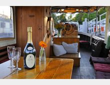 Innenraum eines Bootes mit Holzmöbeln, einer Tischfläche im Vordergrund, auf der eine Champagnerflasche, zwei Gläser und eine Vase mit orangefarbener Blume stehen. Im Hintergrund sind Sitzkissen, ein Steuerrad und Dekorationen zu sehen, mit Blick auf grüne Bäume und eine Hafenkulisse.