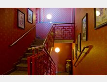 Treppenhaus mit roten Wänden und einem asiatisch inspirierten Geländer in geometrischer Musterung, dekoriert mit eingerahmten Bildern und einer großen Vase auf einer unteren Ebene. Die Wände sind mit goldenem Muster und punktförmiger Beleuchtung ausgestattet.