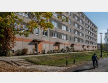 Wohnblock aus Plattenbau mit mehreren Stockwerken, grauer Fassade und roten Ziegeldetails; davor ein schmaler Weg, einige Bäume und ein Kind auf einem Fahrrad.