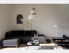 Helles Wohnzimmer mit einer dunkelgrauen Eckcouch, einem schwarzen Beistelltisch und minimalistischer Dekoration. An der weißen Wand hängen zwei gerahmte Kunstwerke, und eine moderne weiße Deckenlampe sowie eine Stehlampe sorgen für Beleuchtung.