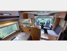 Innenraum eines modernen Wohnmobils mit Holzelementen. Sichtbar sind eine kleine Küchenzeile mit rundem Spülbecken, Sitzgelegenheiten, Schränke, ein Panoramafenster sowie der Fahrerbereich mit zwei drehbaren Sitzen.