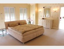 Schlafzimmer mit einem großen beige gepolsterten Bett, zwei Fenstern mit Rollläden, einem runden Beistelltisch und einem weißen Sideboard mit Spiegel darüber. Die Wände sind mit cremefarben-tapeten mit dezentem Muster dekoriert, und ein Kronleuchter hängt von der Decke.