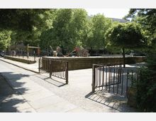 Städtischer Platz mit einem umzäunten Bereich, in dem sich ein kleiner Park mit Bäumen und einem Spielplatz befindet. Im Hintergrund stehen Gebäude und geparkte Fahrzeuge entlang einer Straße.