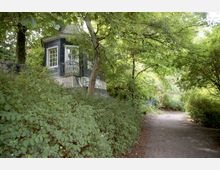 Kleines, dunkelblaues Pavillon-Gebäude mit weißen Fenstern und einem dekorativen Geländer, umgeben von dichtem Grün und Bäumen; im Vordergrund ein schmaler, unbefestigter Weg.