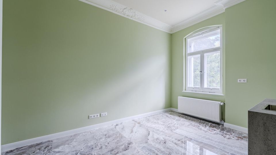 Heller, leerer Innenraum mit hellgrünen Wänden, weißer Stuckdecke und glänzendem Marmorboden. An der Stirnseite befindet sich ein großes Rundbogenfenster über einem Heizkörper; rechts ist eine graue Arbeitsplatte mit eingelassenem Spülbecken angeschnitten.