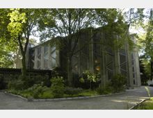Außenansicht einer modernen Kirche mit geometrisch gestalteten Glasfenstern und Betonwänden, umgeben von einem Garten mit Bäumen und Sträuchern. Ein gepflasterter Weg führt an der Kirche vorbei.