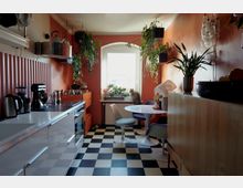 Kleine Küche mit orangefarbenen Wänden, schwarz-weiß kariertem Boden und hängenden Zimmerpflanzen. Auf der linken Seite befinden sich eine weiße Arbeitsplatte mit Küchengeräten, rechts ein Esstisch mit modernen Stühlen vor einem Fenster, auf dessen Fensterbank Pflanzen und eine Katze sitzen.