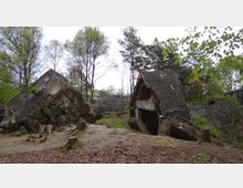 Zerfallene Betonruinen inmitten eines bewaldeten Gebiets mit jungen und ausgewachsenen Bäumen; die zerstörten Strukturen zeigen massive Betonblöcke und Risse, die auf historische Schäden hinweisen. Der Boden ist mit Laub bedeckt, und vereinzelt sind Baumstümpfe zu sehen.