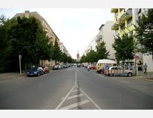 Straßenansicht in einer Stadt mit mehrstöckigen Wohnhäusern auf beiden Seiten, flankiert von Bäumen entlang der Gehwege. Im Hintergrund ragt ein Kirchturm mit grünem Dach hervor, während Autos entlang der Straße und am Straßenrand parken.