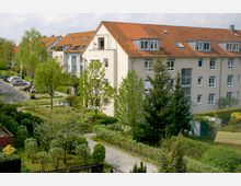 Mehrfamilienhäuser mit cremefarbenen Fassaden und roten Dächern in einer grünen Wohngegend. Im Vordergrund sind gepflegte Gärten, Gehwege und Bäume zu sehen, während parkende Autos entlang einer Straße im Hintergrund sichtbar sind.