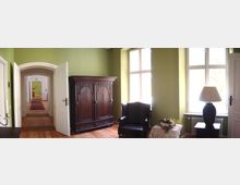 Innenraum eines historischen Hauses mit Parkettboden und grünen Wänden. Zu sehen sind ein antiker Holzschrank, ein schwarzer Polstersessel, ein Tisch mit einer Lampe und Blumendekoration sowie ein Flur mit offenen Türen und einem Teppichläufer.