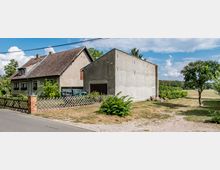 Zwei Wohnhäuser auf einem ländlichen Grundstück, eines mit Ziegeldach und sichtbarem Dachfenster, das andere mit unverputzter Betonfassade. Im Vordergrund ein niedriger Holzzaun, dahinter geparkte Fahrzeuge und umgebende Grünflächen mit Bäumen.