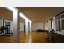 Großer, offener Raum mit Parkettboden, weißen Säulen und moderner Beleuchtung. Im Hintergrund befinden sich eine Sitzgruppe mit Sofa und Stehlampe, rechts ein langer Holztisch mit Hockern, links eine Glastür und Grünpflanze.