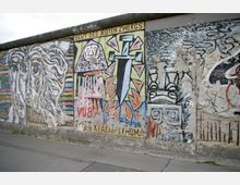 Bemalte Betonwand, Teil der Berliner Mauer, mit auffälligen Graffiti und Kunstwerken, darunter bunte, abstrakte Figuren und Schriftzüge. Die Bilder sind teils verwittert, mit sichtbaren Zeichen der Zeit.