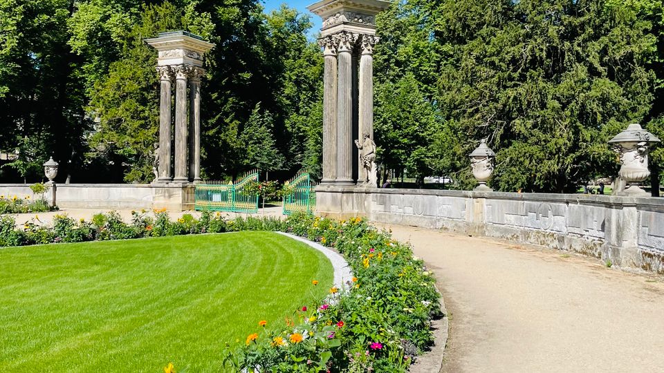 Parkanlage mit geschwungenem Kiesweg entlang eines Blumenbeets und einer großen Rasenfläche. Im Hintergrund stehen zwei hohe steinerne Säulenmonumente neben einer niedrigen Balustrade, umgeben von dichtem Baumbestand unter blauem Himmel.