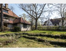 Ein Garten mit einer Wiese und mehreren hohen Bäumen, im Hintergrund ein Haus mit rötlichem Ziegeldach und verputzter Fassade. Rechts sind weitere Häuser mit ähnlichem Baustil zu sehen, umgeben von Hecken und Bäumen.