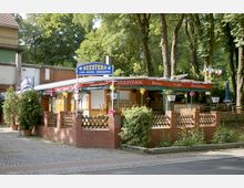 Straßenseitige Ansicht eines Cafés und Biergartens mit dem Namen "Seestern", umgeben von hohen Bäumen. Das Gebäude ist teilweise aus Holz, mit einem roten Markisendach und einer niedrigen Ziegelmauer eingefasst, dekoriert mit Pflanzen und bunten Lichtern.