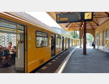 Ein gelber U-Bahn-Zug hält an einem überdachten Bahnsteig mit einem hölzernen Dach und gewölbten Fenstern. Eine digitale Anzeigetafel zeigt Abfahrtsinformationen, und eine Uhr ist am Pfeiler montiert; Passagiere sitzen im Zug oder stehen am Bahnsteig.