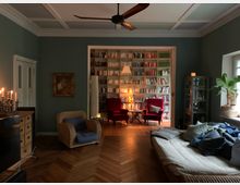 Wohnzimmer mit dunkelgrünen Wänden und Holzboden; im Hintergrund ein Bücherregal, flankiert von zwei roten Sesseln mit Beistelltischen und einer Stehlampe. Im Vordergrund ein Sofa mit Kissen, ein Rattansessel sowie eine Kommode mit Kerzenleuchtern. Ein großes Fenster rechts lässt Tageslicht herein.