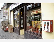 Straßenszene mit einer kleinen Spirituosengeschäft-Fassade, die große Fenster mit ausgestellten Flaschen und Dekorationen zeigt. Daneben stehen Kartons und ein roter, wandmontierter Kaugummiautomat an einer gelben Hauswand.