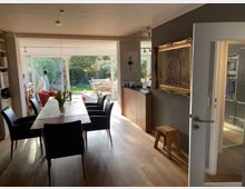 Essbereich in einem modernen Wohnhaus mit Holzfußboden, einem langen Esstisch und sechs schwarzen Stühlen. Große Fensterfront im Hintergrund mit Blick auf einen Garten mit Rasen und Gartenmöbeln; an der rechten Wand ein großes, gerahmtes Gemälde.