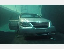 Underwater scene showing a silver minivan inside a submerged tunnel or parking structure, with concrete walls and a ladder visible in the background. The vehicle is partially tilted on the floor, surrounded by murky water and scattered debris.