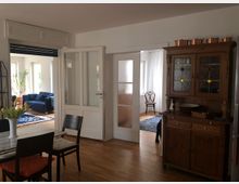 Wohnraum in einem hellen Apartment mit Holzboden, einem alten Holzschrank rechts und Glastüren, die zu zwei weiteren Räumen führen. Im Hintergrund sind ein Wohnzimmer mit Sofa und ein weiterer Bereich mit Stühlen sichtbar.