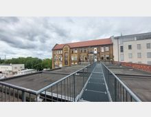 Backsteinbau mit rotem Satteldach im Hintergrund, gesehen über eine flache Dachfläche mit einem Metall-Steg, der von der Kamera zur Gebäudefassade führt. Im Hintergrund sind weitere Gebäude und Bäume unter bewölktem Himmel sichtbar.