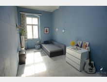 Schlicht eingerichteter Raum mit blauen Wänden, einem einzelnen Bett an der rechten Wand, einer weißen Kommode mit darauf stehenden Dekorationsgegenständen und einer Gitarre daneben. Ein großes Fenster mit gemusterten Vorhängen lässt Tageslicht herein, und eine Zimmerpflanze steht links im Raum.