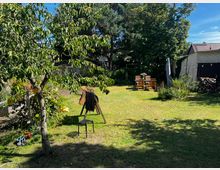 Grüne Gartenlandschaft mit Rasenfläche, einem kleinen Baum im Vordergrund und Blumenbeeten im Hintergrund. Im hinteren Bereich stehen ein Holz-Esstisch mit Stühlen und eine Stehlampe, daneben eine Garage sowie Fahrräder. Ein kindergroßes Holzspielzeug in Form eines Pferdes befindet sich links vorne.