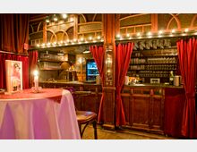 Gemütliche Bar mit rustikalen Holzmöbeln, roten Samtvorhängen und dekorativer Beleuchtung. Im Hintergrund sind Gläserregale, ein Tresen und eine Kühleinheit mit Flaschen sichtbar, im Vordergrund ein Bistro-Tisch mit Kerze.