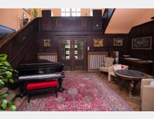 Ein stilvoll eingerichtetes Wohnzimmer mit dunkler Holzvertäfelung, einem schwarzen Flügel mit rotem Hocker, einem großen, roten Orientteppich und antiken Möbeln. An den Wänden hängen Gemälde in goldenen Rahmen, und im Hintergrund befindet sich eine zweiflügelige Holztür mit Glaseinsatz.