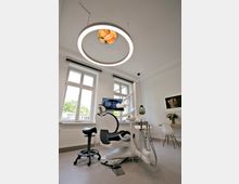 Moderne Zahnarztpraxis mit einem Behandlungsstuhl, einer Zahnärztelampe und angeschlossenen Geräten. Im Hintergrund sind große Fenster, ein Schreibtisch mit Blumen und ein Röntgenbild auf einem Bildschirm sichtbar.