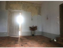 Ein schlichter Innenraum mit einem Holztürdurchgang, durch den helles Licht von einem angrenzenden Raum fällt. Der Boden ist aus rustikalen Terrakottafliesen, an der Wand hängt verblasste Stuckverzierung, darunter ein Blumenarrangement in einer Vase; rechts führen Holzstufen nach oben.