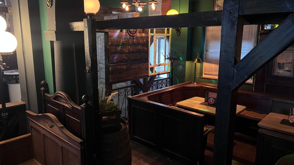 Gemütlicher Pub-Innenraum mit dunklen Holztrennwänden und Sitznischen, Holzbänken und Tischen sowie grünen Wänden mit gerahmten Schildern. Warme Hängelampen, ein Fenster mit Jalousien und dekorative Elemente wie Geländer und Fass ergänzen die rustikale Atmosphäre.
