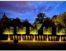 Nachtszene eines Gartens mit gleichmäßig geschnittenen Bäumen, die von unten durch warmes Licht angestrahlt werden, und hohen Ziergräsern im Hintergrund. Im oberen Teil sind Bäume und ein Gebäude mit Giebeldach vor einem wolkigen, dunkelblauen Himmel sichtbar.