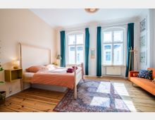 Helles Schlafzimmer mit Holzfußboden, einem Doppelbett mit orangefarbener Bettwäsche, großen Fenstern mit türkisfarbenen Vorhängen, einem Teppich im orientalischen Stil, einer orangen Couch und dekorativen Pflanzen. Die Wände sind hellbeige, und an einer Wand hängen gerahmte Bilder.