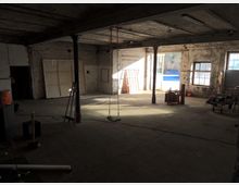 Industriehalle mit offenliegender Betonkonstruktion, brüchigen Backsteinwänden und mehreren Stahlsäulen, die den Raum stützen. Tageslicht fällt durch große Fenster und ein offenes Tor ein, im Raum befinden sich verschiedene Werkzeuge und Gegenstände, darunter eine Schaukel.